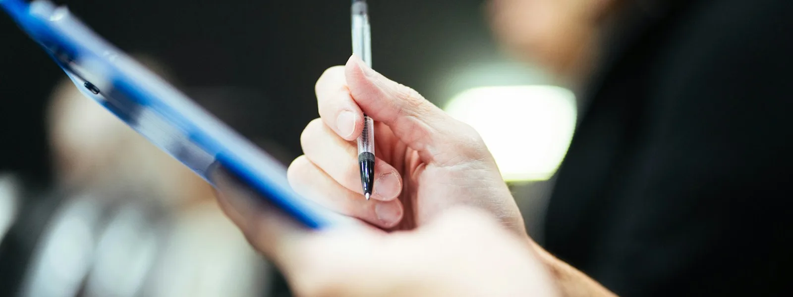 Une main tient un stylo au-dessus d'une planchette porte-formulaire bleue